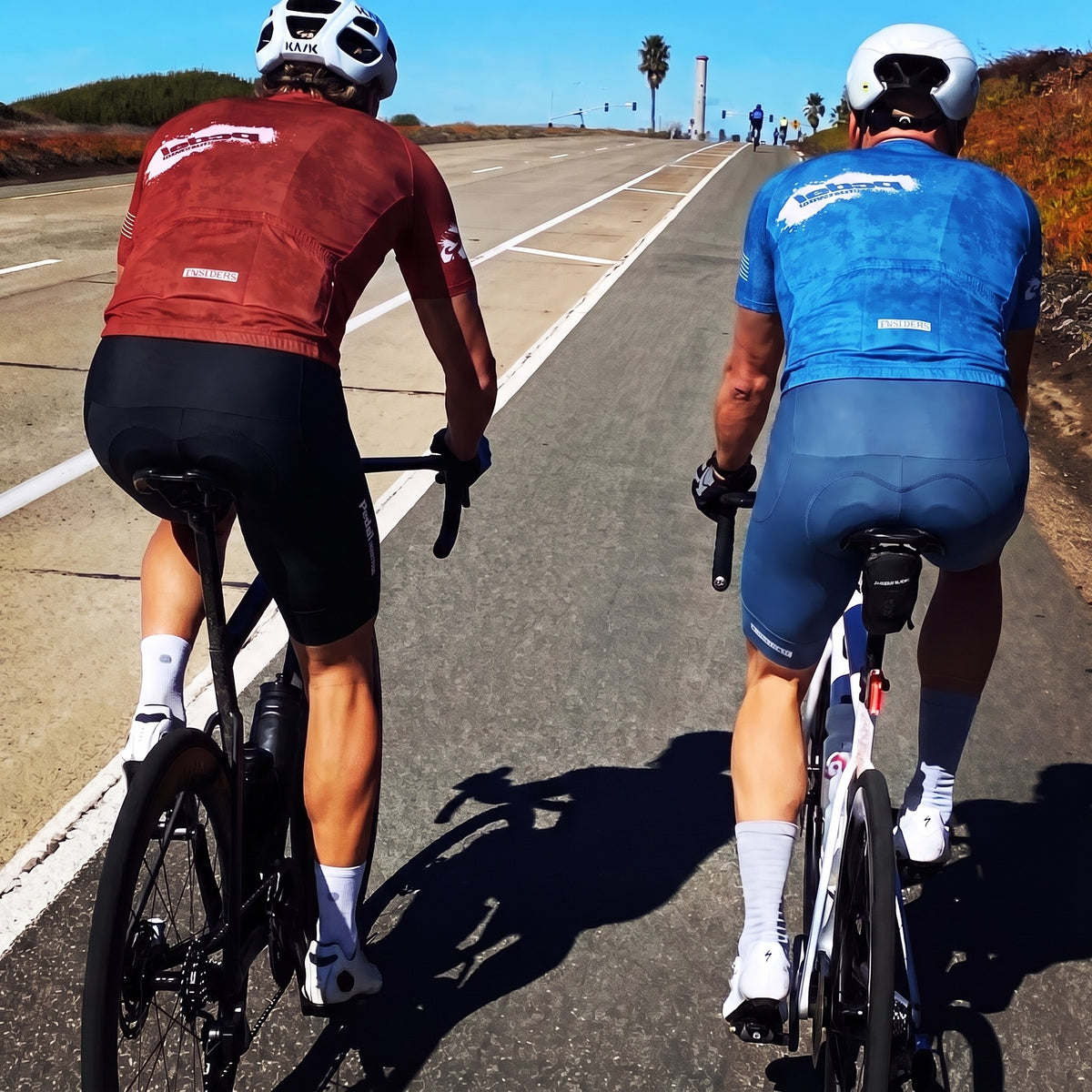 Two riders in PEDAL Insiders 2027 kits, red and azure blue, seen from behind on a coastal bike lane