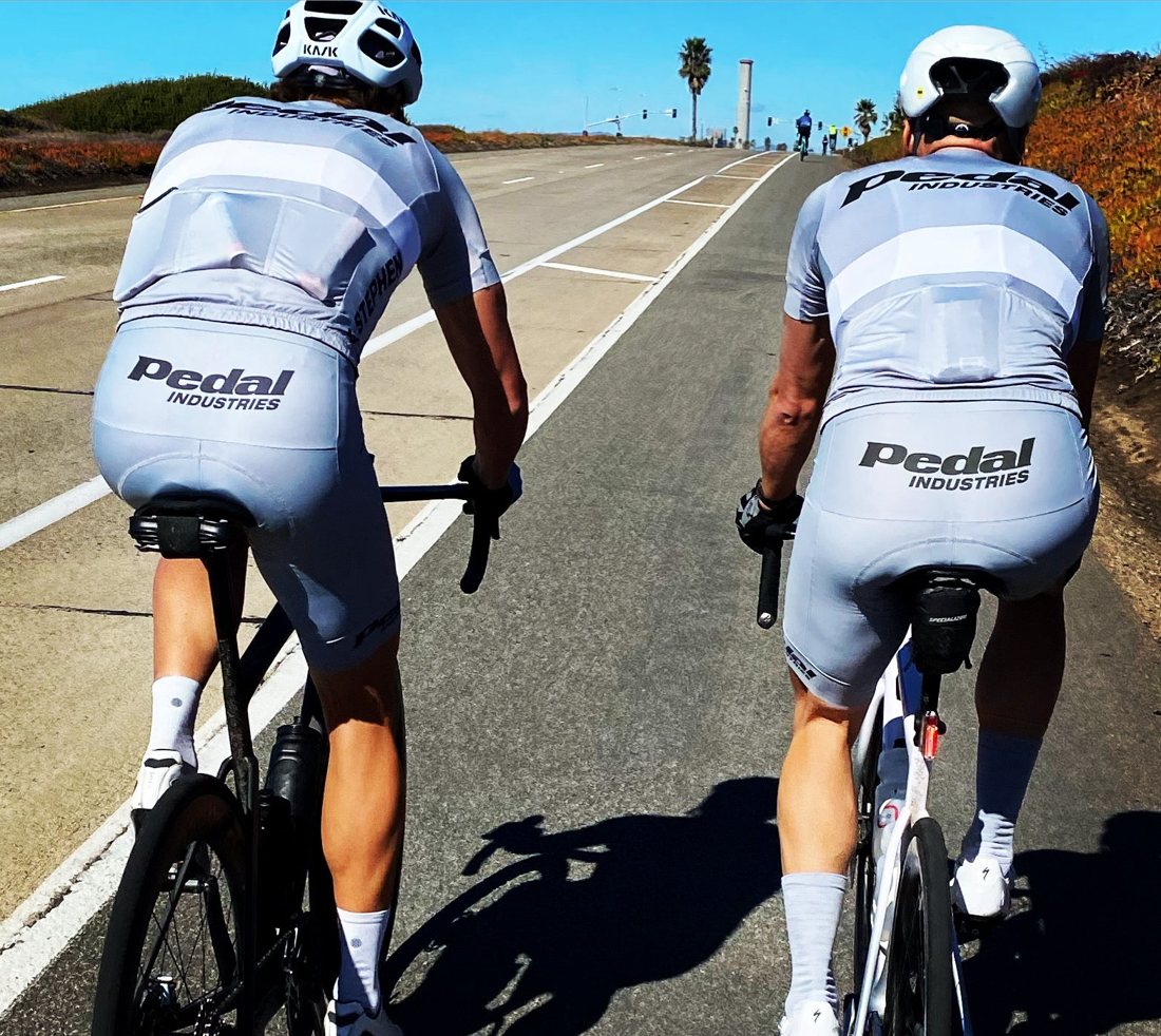 Two riders in matching Pedal jerseys and bibs on a coastal road